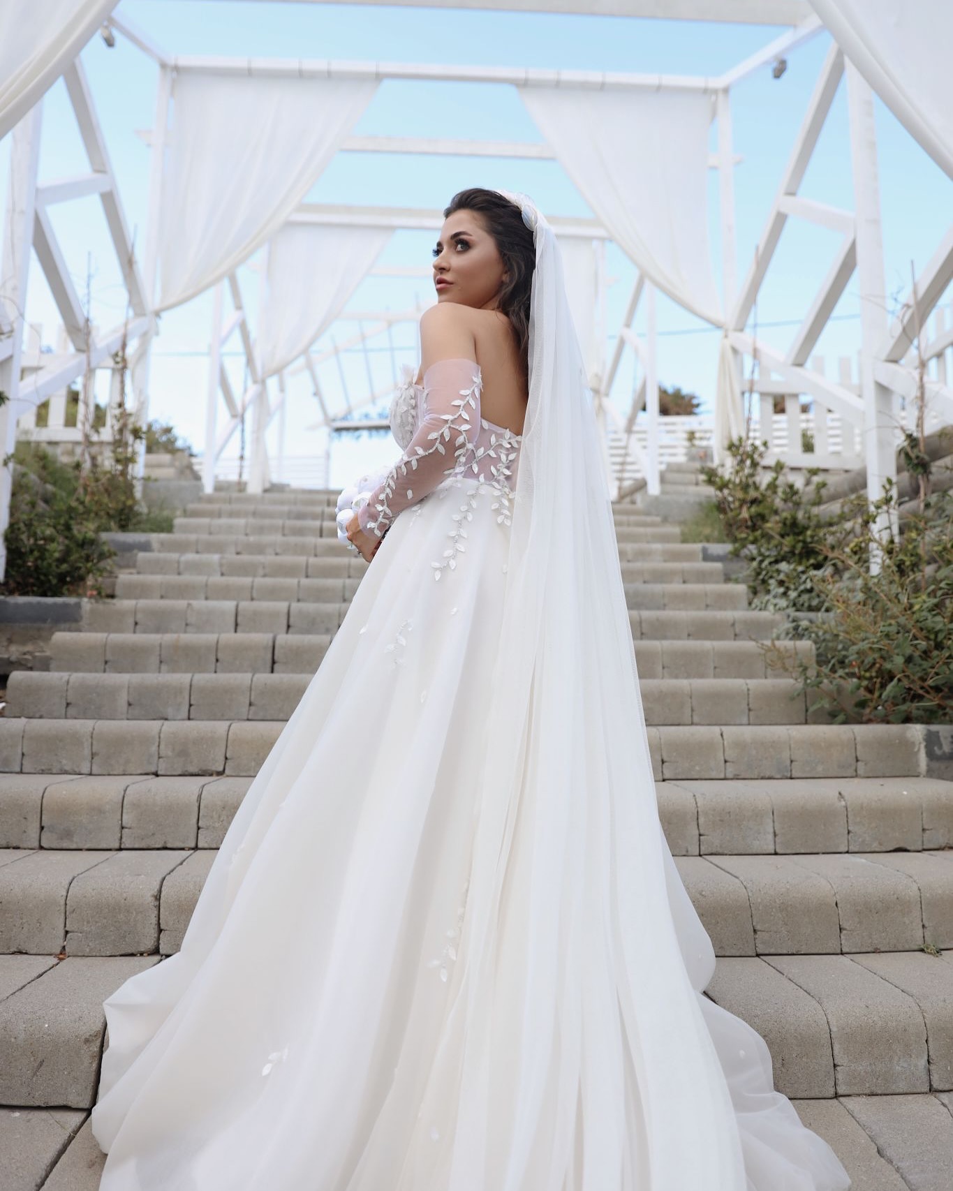 Off-shoulder transparan çiçek işlemeli kollara sahip A kesim gelinlik, uzun duvak detayıyla merdivenlerde arkadan görünüm.