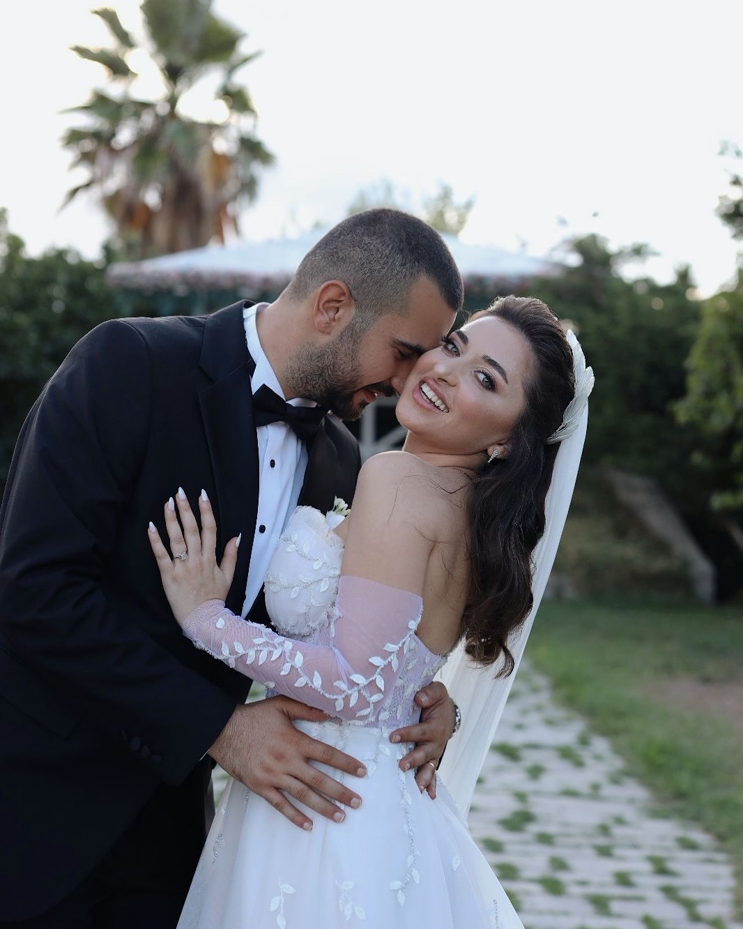 Romantik çiçek aplikeli off-shoulder korse gelinlik ile damatla sarılma pozu, zarif duvak ve A kesim etek görünümü.