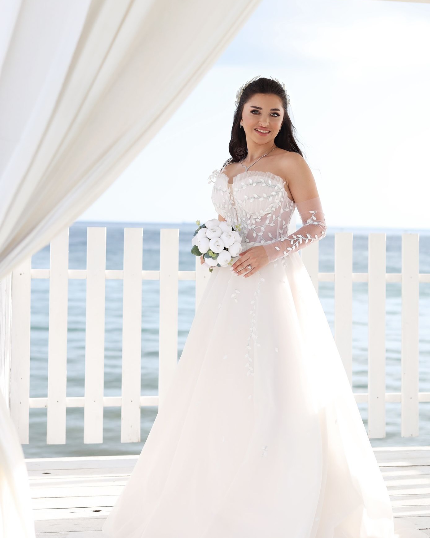 Deniz manzarası önünde Aliko Couture off-shoulder A kesim gelinlik, çiçek işlemeli transparan kollar ve sade buketle tam boy çekim.