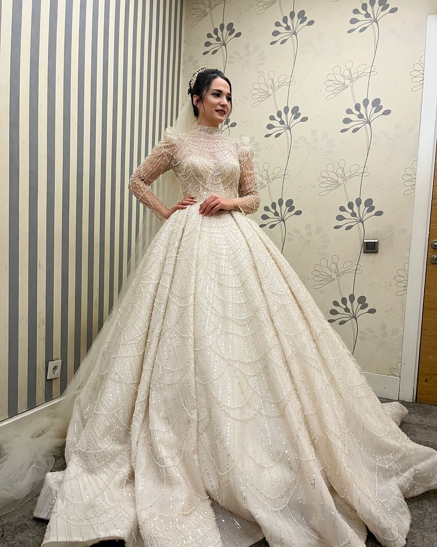 Yüksek yakalı uzun kollu taş işlemeli prenses gelinlik, bel vurgulu ışıltılı kumaş detayıyla iç mekân çekimi