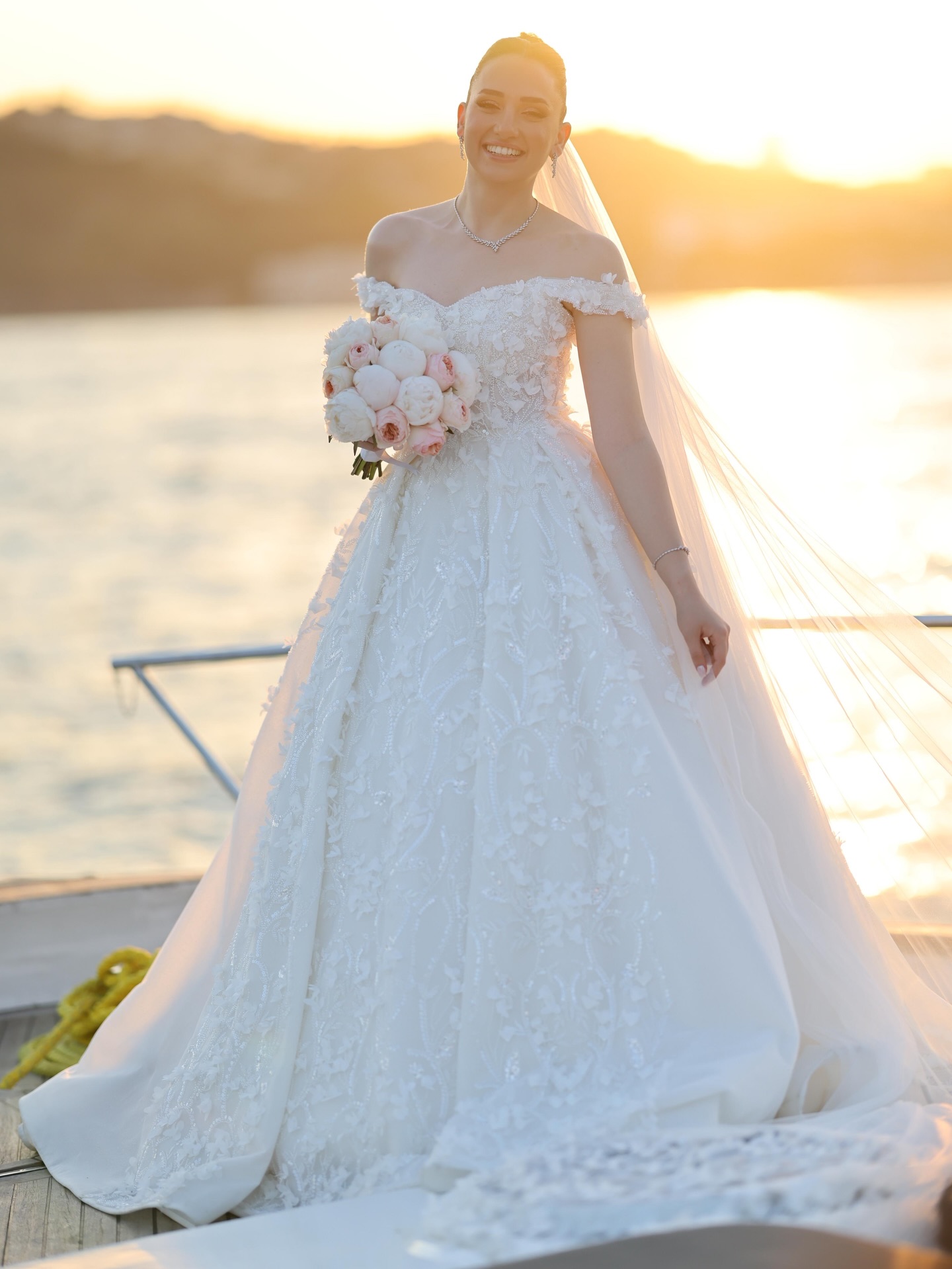 “Ceylan Güres Modaevi düşük omuz prenses gelinlik modeli, gün batımı ışığında ışıltılı etek dokusu ve romantik çiçek işlemeleriyle tam boy görünüm”