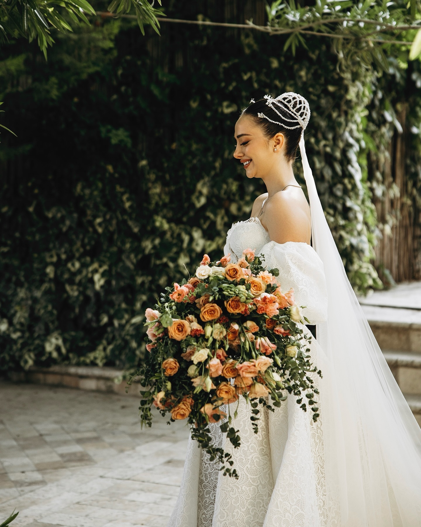 Taç ve duvak detaylı, omuzdan düşük dantel A kesim gelinlik ile bahçede gelin poz veren gelin