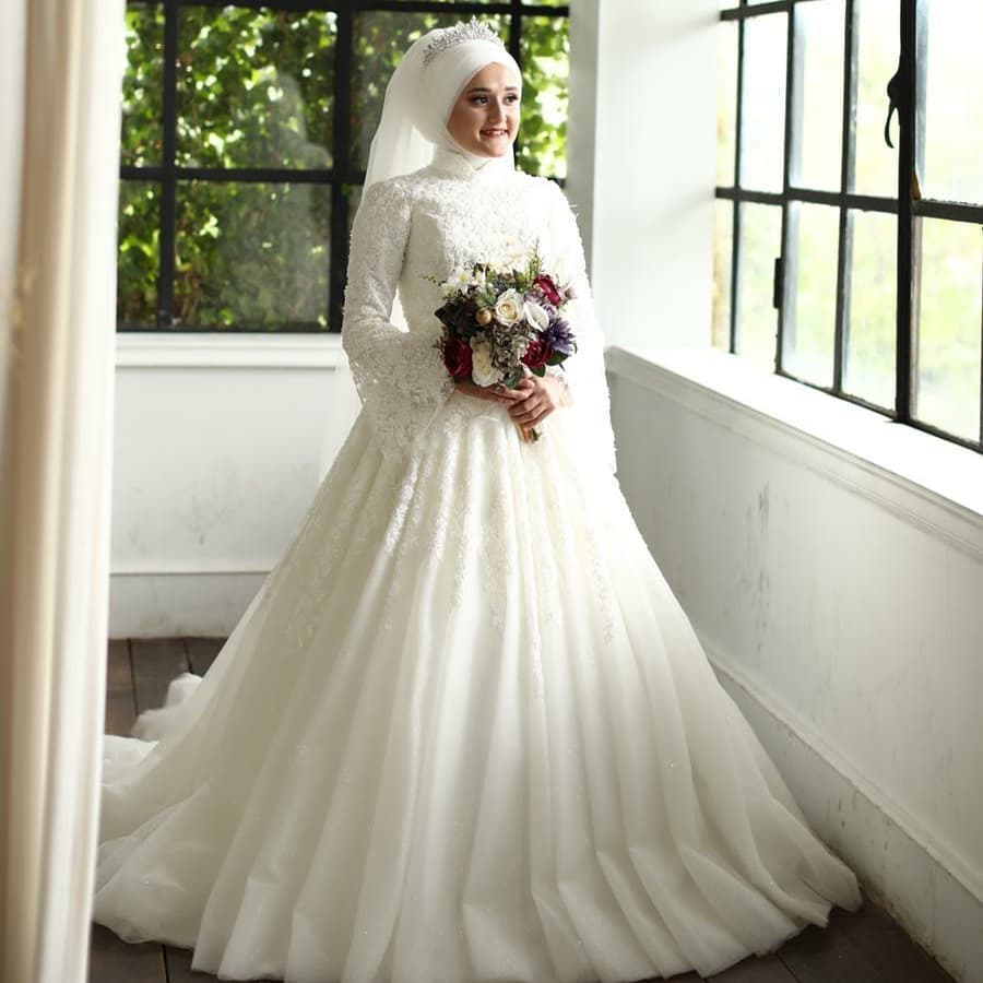 Deniz Moda Evi tesettür prenses gelinlik modeli, uzun kollu yüksek yakalı işlemeli üst beden ve kabarık A kesim etek tasarımıyla iç mekanda gelin pozu.