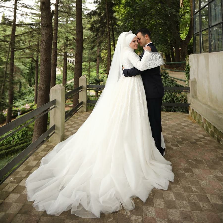 Deniz Moda Evi tesettür gelinlik modeli, uzun duvak ve kuyruk detaylı prenses siluet ile dış mekanda romantik çift pozu.