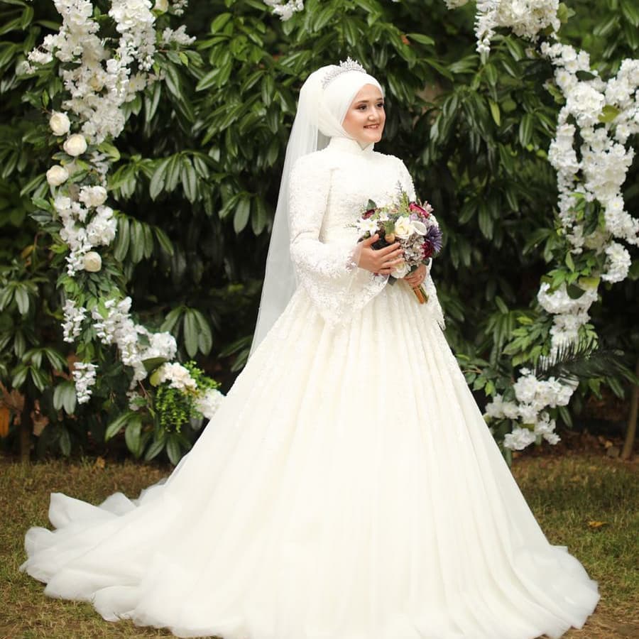 Deniz Moda Evi tesettür prenses gelinlik, ışıltılı dokulu kabarık etek ve işlemeli üst beden detayıyla çiçekli dış çekim konseptinde gelin görünümü.