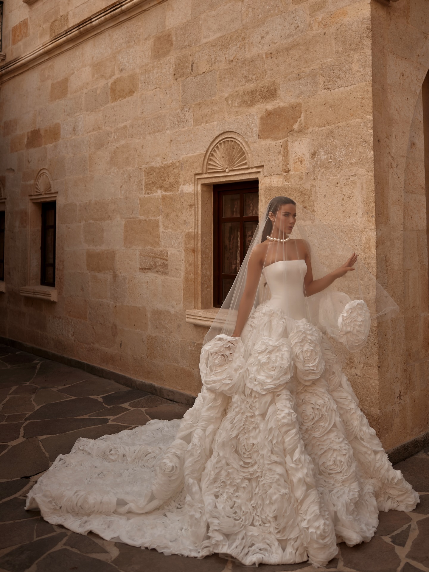 Straplez prenses kesim gelinlik, 3D çiçek dokulu kabarık etek ve uzun kuyruk detayıyla taş duvar önünde dış çekim.
