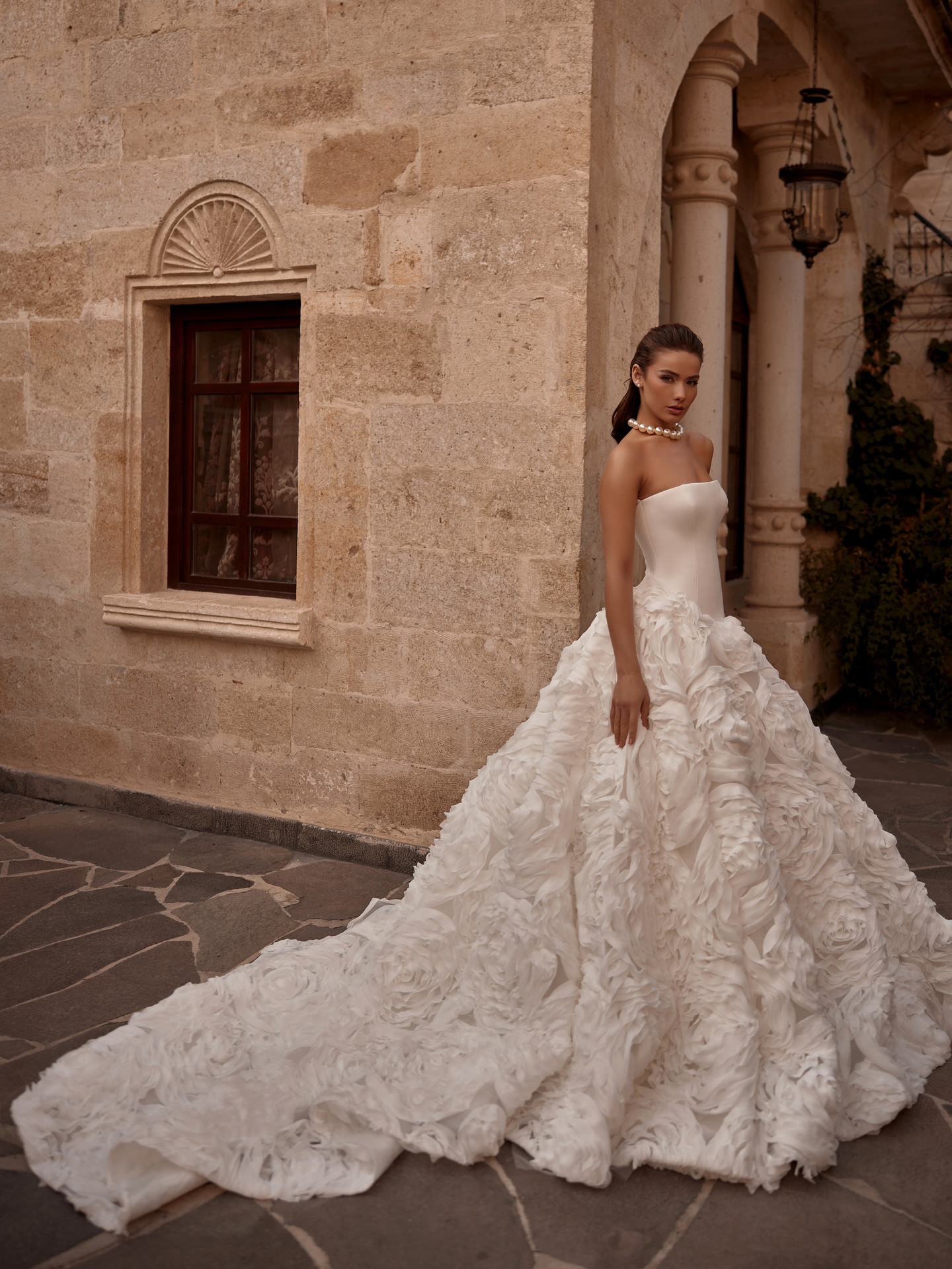 Minimal straplez üst beden ve hacimli çiçek aplikeli etek tasarımına sahip romantik prenses gelinlik, tarihi mekanda poz veriyor.
