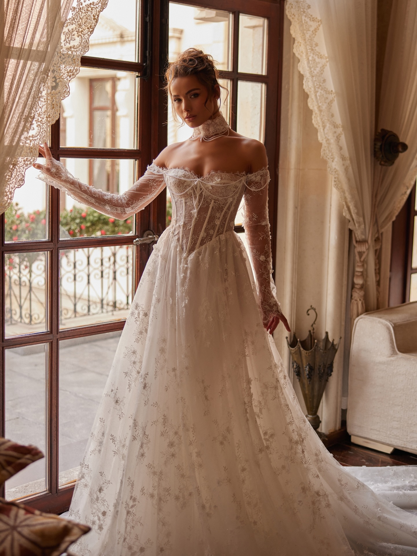 Düşük omuzlu, dantel uzun kollu ve korseli üst tasarıma sahip A kesim prenses gelinlik ile pencere önünde zarif gelin pozu.