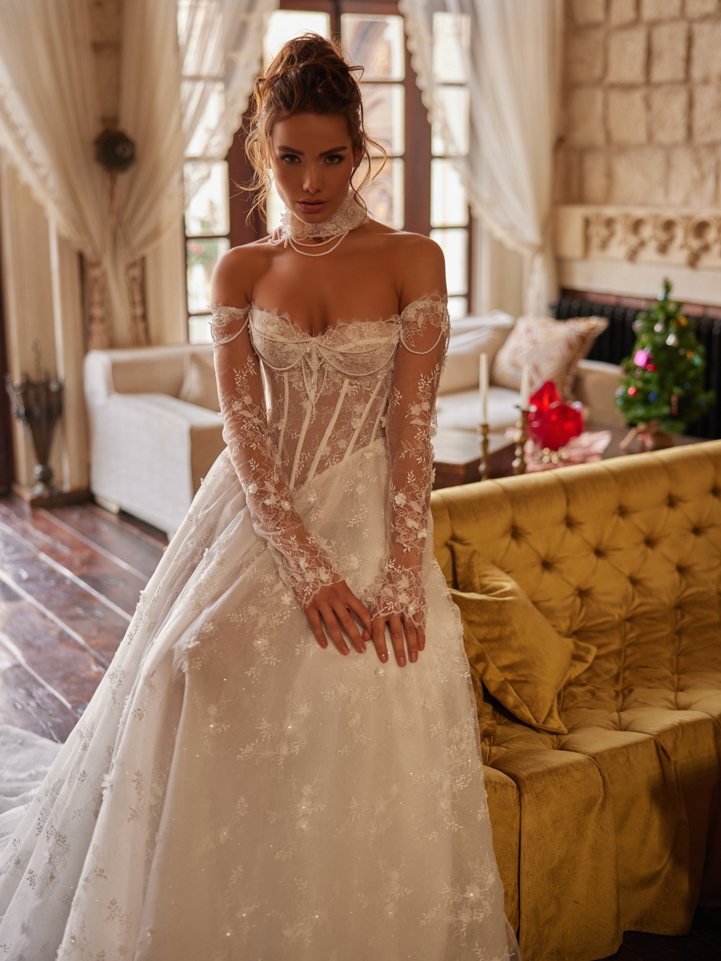 Dantel kollar ve korseli üst tasarımıyla öne çıkan düşük omuz gelinlik modeli, sıcak tonlu iç mekanda couture gelin duruşu.