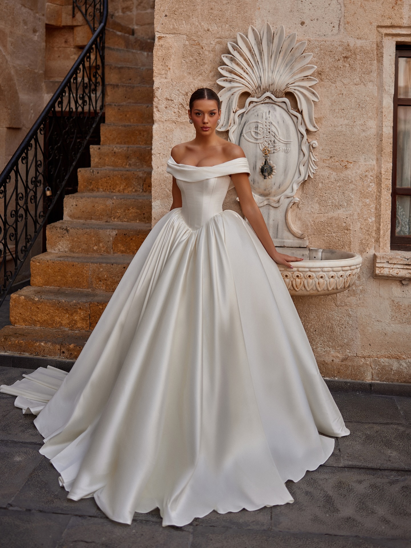 Taş dokulu mekanda çekilmiş, hacimli etekli prenses siluet saten gelinlik tam boy görünüm.