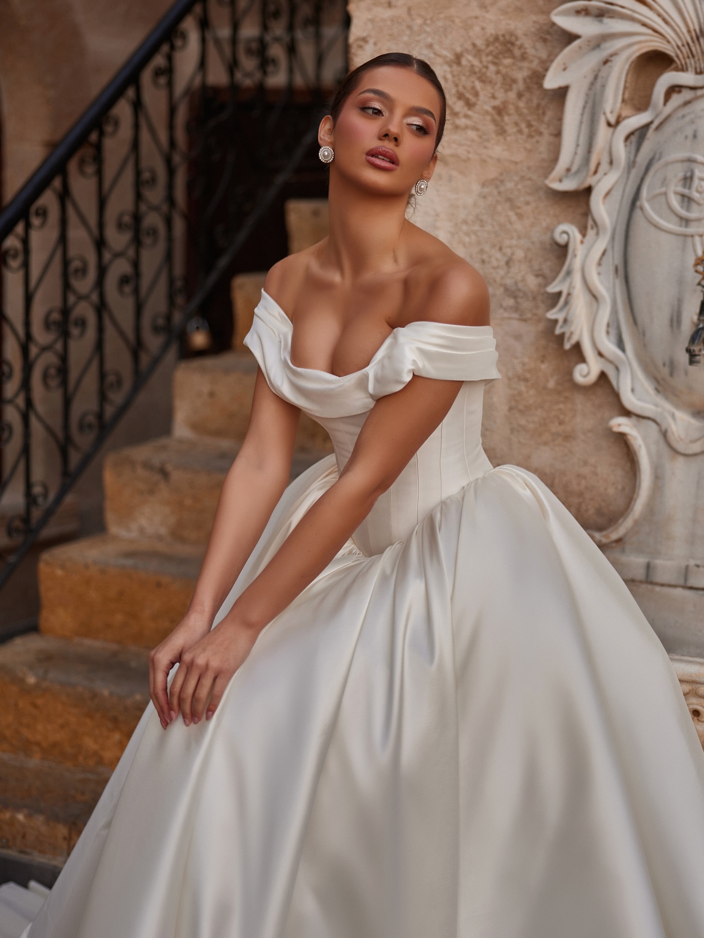 Düşük omuz prenses gelinlik, parlak saten kumaş ve güçlü siluetle oturarak poz.