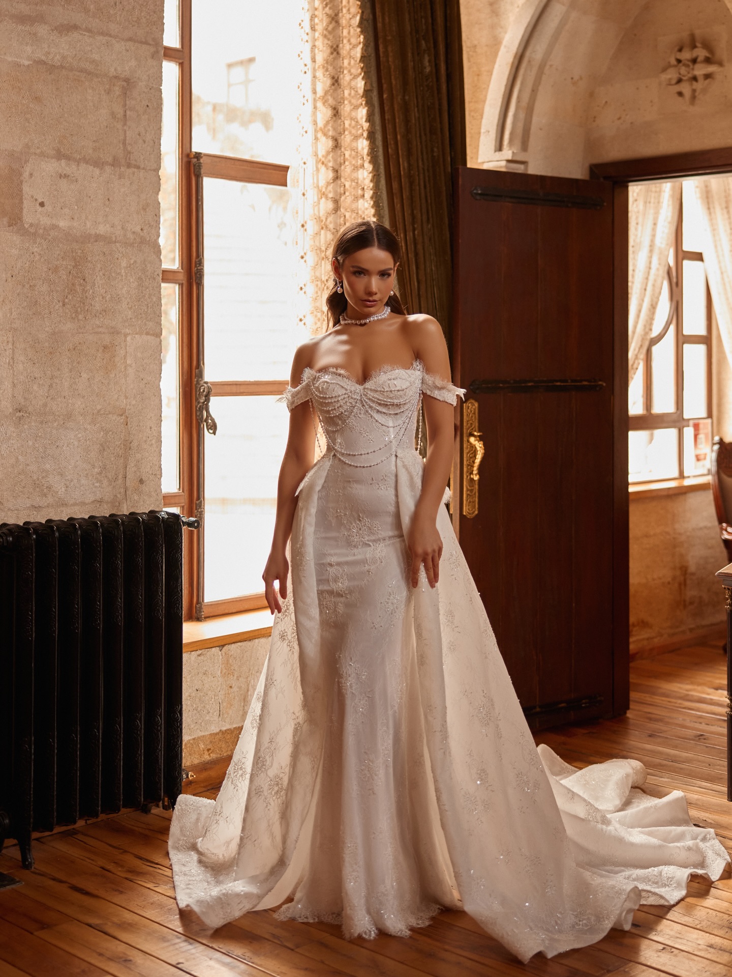 “İnci drapeli düşük omuz balık gelinlik, uzun kuyruk detayıyla tam boy çekimde asil duruş.”