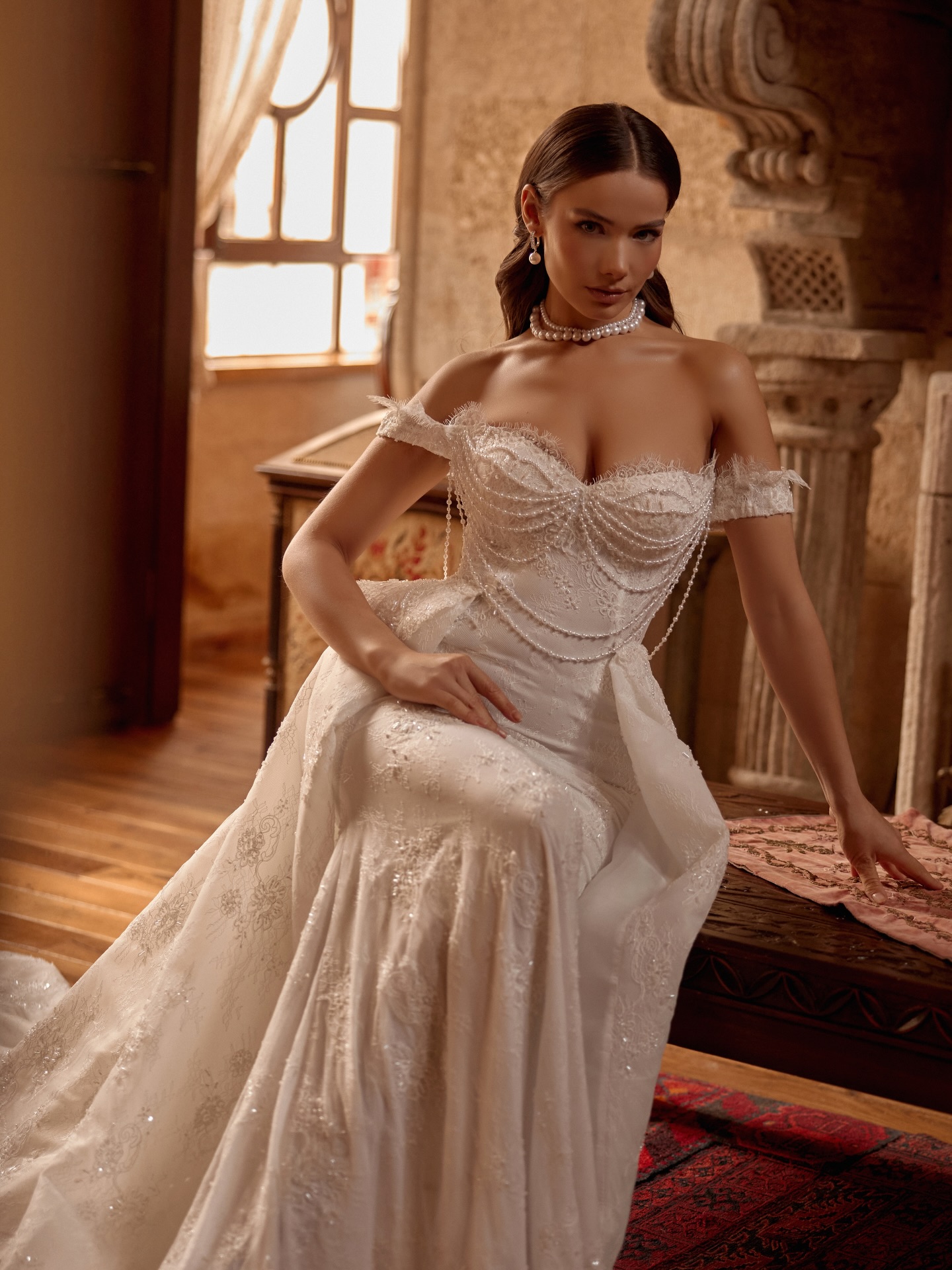 “İnci detaylı düşük omuz balık gelinlik, oturuş pozunda dantel işlemeler ve şık korsaj yapısıyla öne çıkıyor.”