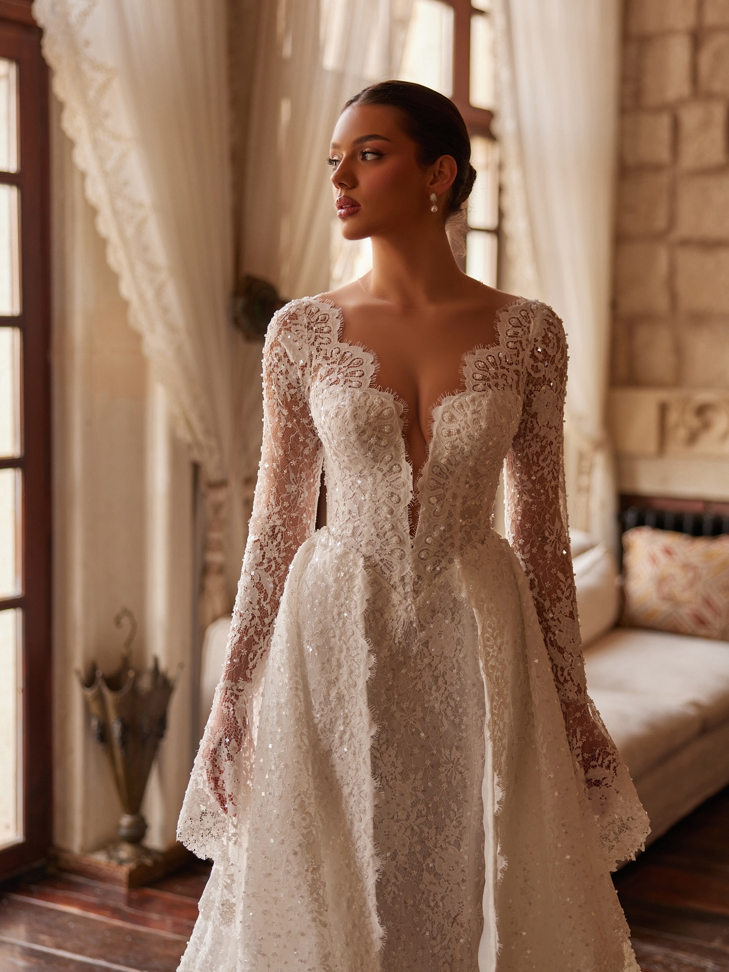 Yumuşak ışıkta uzun kollu dantel gelinlik, derin V yaka ve belden açılan A kesim form ile zarif tasarım.