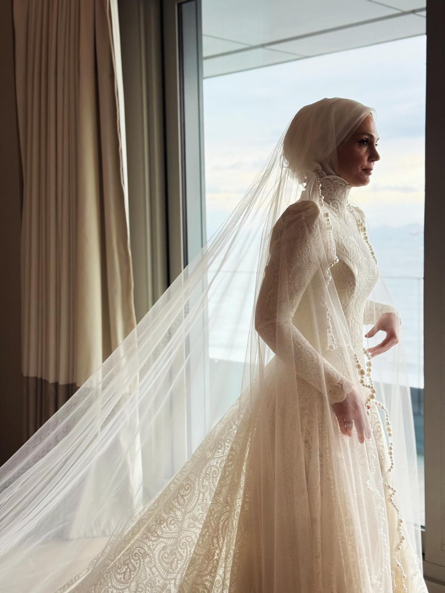 “Özem Keskin yüksek yaka dantelli tesettür gelinlik, uzun duvak ve romantik A kesim siluet – pencere önü gelin pozu”