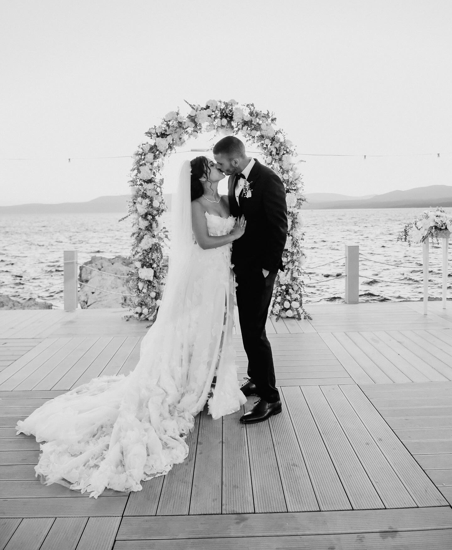 Çiçek kemer önünde öpüşme anı, uzun duvak ve kabartmalı gelinlik dokusu (siyah beyaz)