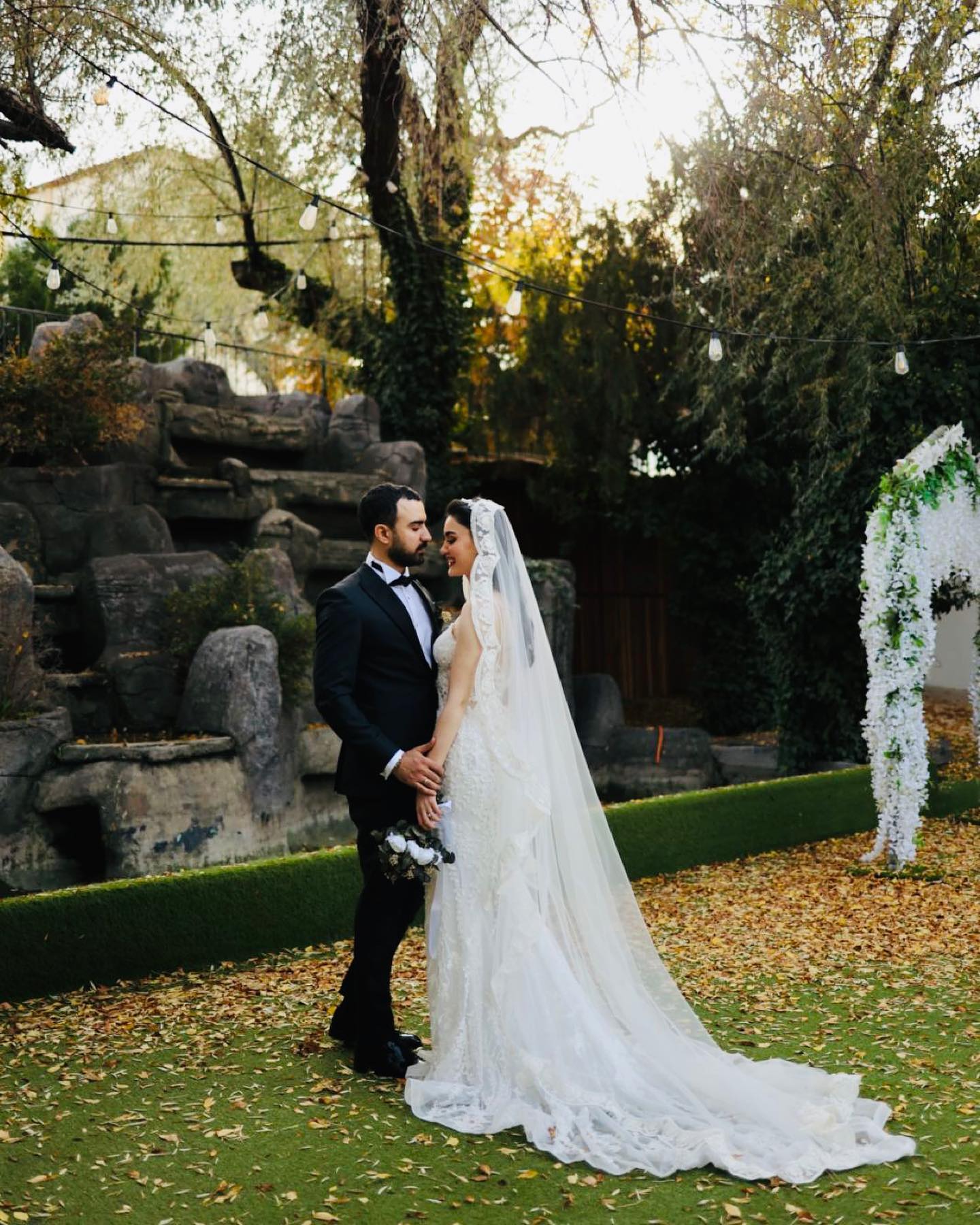 Bahçede romantik çekim, uzun duvaklı dantel gelinlik ve damat – sonbahar yaprakları