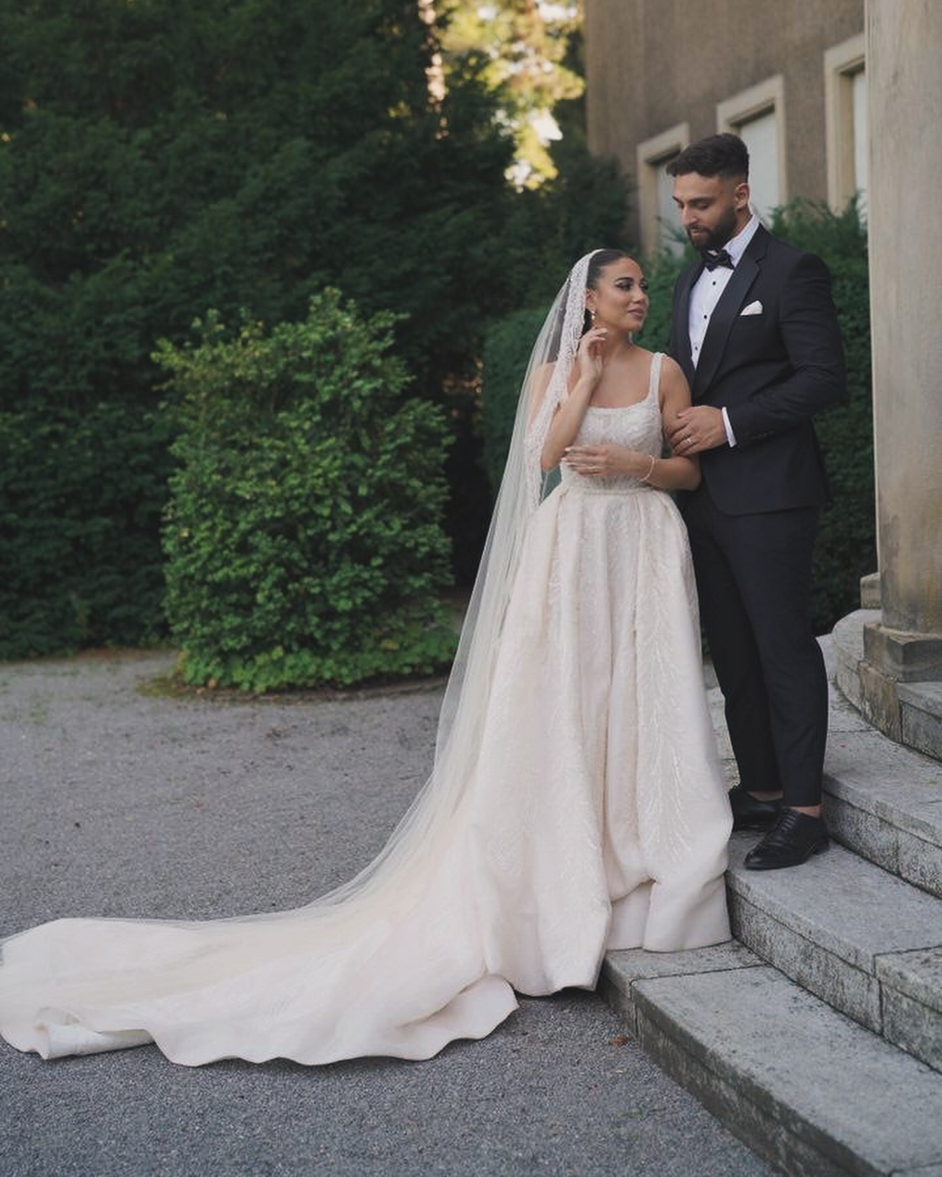 “İnce askılı kare yaka, ışıltılı dokulu A kesim prenses gelinlik ve uzun kuyruk detaylı dış çekim pozu.”