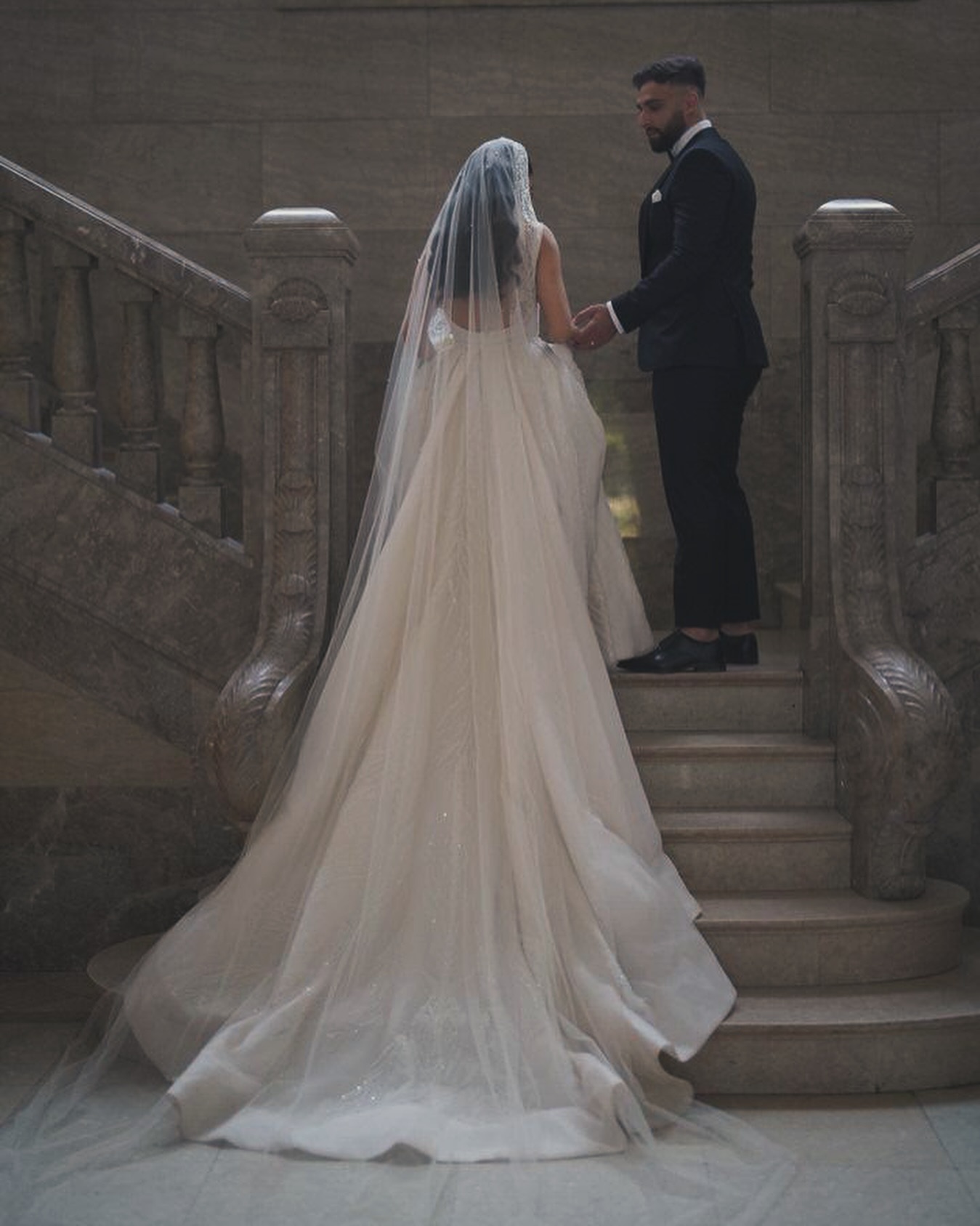 “Merdiven başında uzun kuyruklu prenses gelinlik ve duvakla çekilmiş romantik ve dramatik dış çekim karesi.”