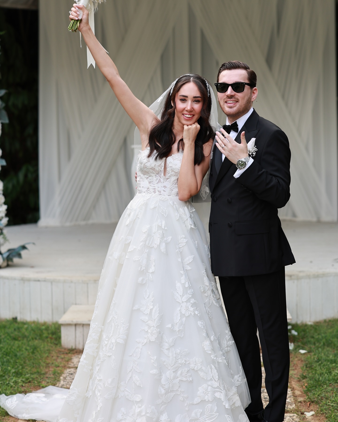  Çiçek işlemeli A kesim gelinlik giyen gelin ve smokinli damadın dış mekânda el ele poz verdiği düğün fotoğrafı.