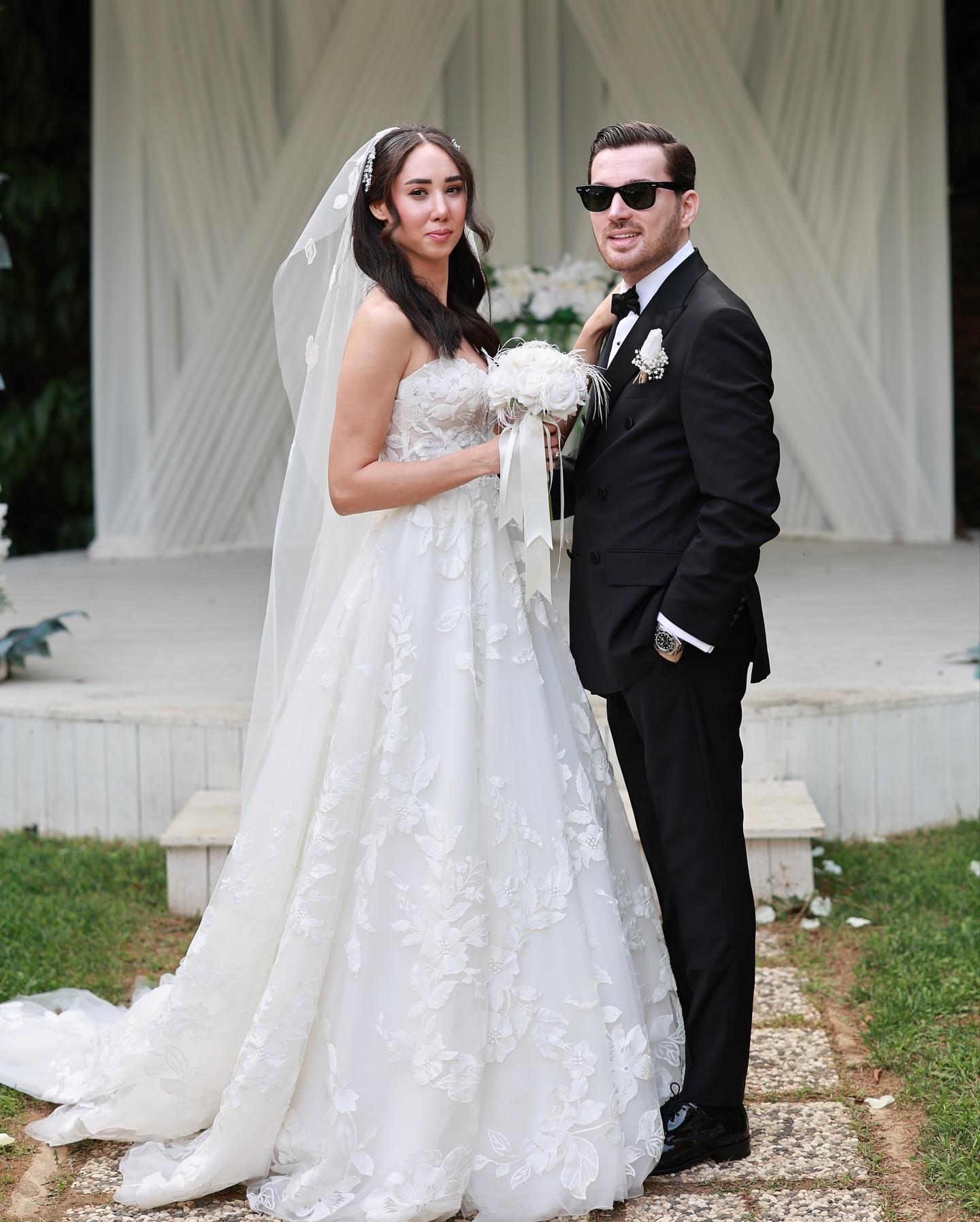 Çiçek detaylı A kesim gelinlik ve uzun duvakla gelin ile smokinli damadın dış mekânda şık poz verdiği düğün karesi.