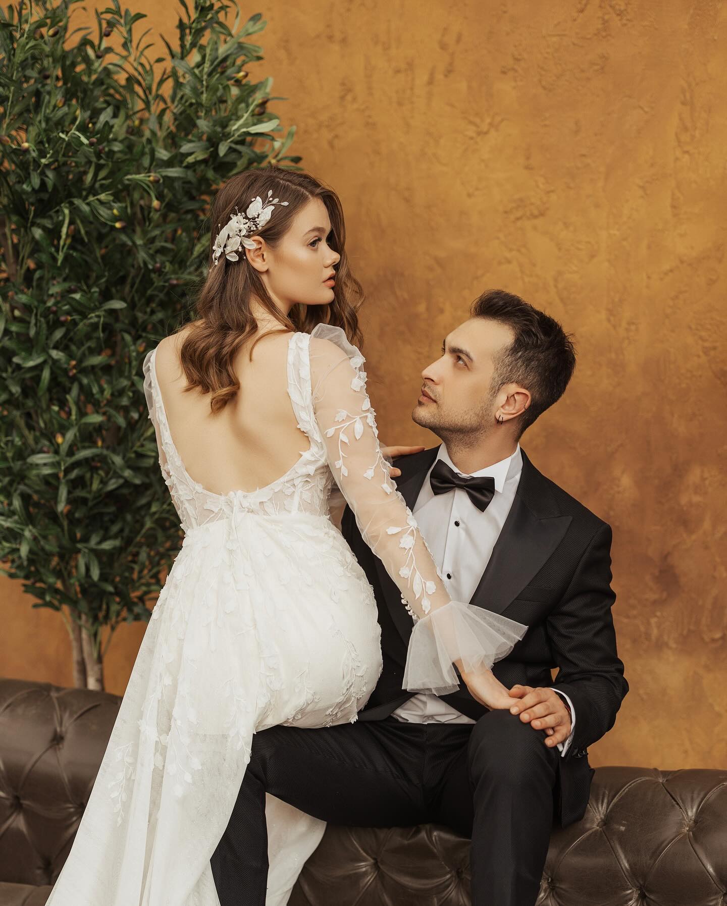 Derin sırt dekolteli, çiçek işlemeli uzun kollu A kesim gelinlik ile damat yanında romantik poz