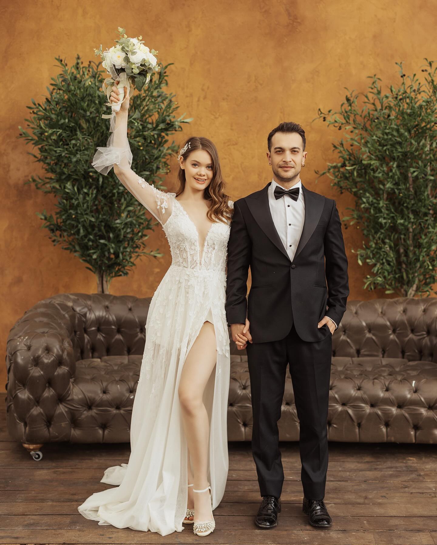 Derin V yakalı, yırtmaçlı ve 3D çiçek detaylı gelinlik giyen gelin ile damadın tam boy düğün pozu.